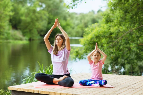 Mom and daughter perform a yoga exercise on the river bank on a warm summer day. Healthy lifestyle concept