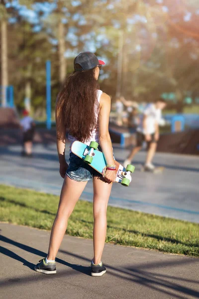 Young brown-haired woman with a skateboard in her hands on the background of a skateboarding platform. The model stands with her back