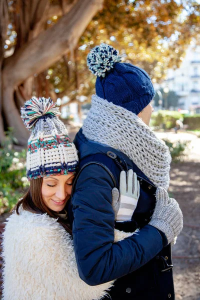 Young woman dressed in winter gloves and woolen hat hugs in love and with eyes closed to her boyfriend from behind