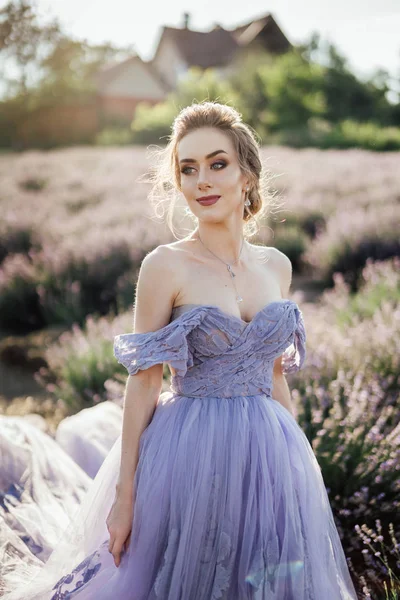 beautiful young woman in purple dress posing on lavender field with houses on background