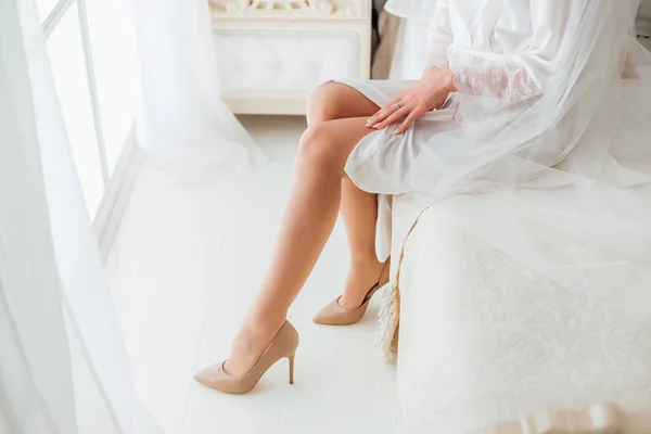 新娘穿着精致的柔和白色连衣裙和优雅的鞋子坐在房间里的床上 — 图库照片