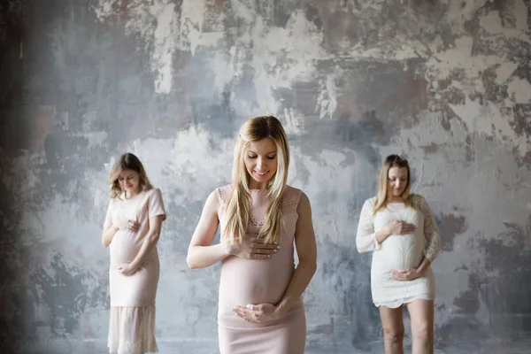 Três mulheres grávidas ficam de pé com sorrisos felizes e olham para a barriga.Uma mulher grávida em foco e duas mulheres grávidas em um desfoque. Fundo traseiro parede cinza — Fotografia de Stock