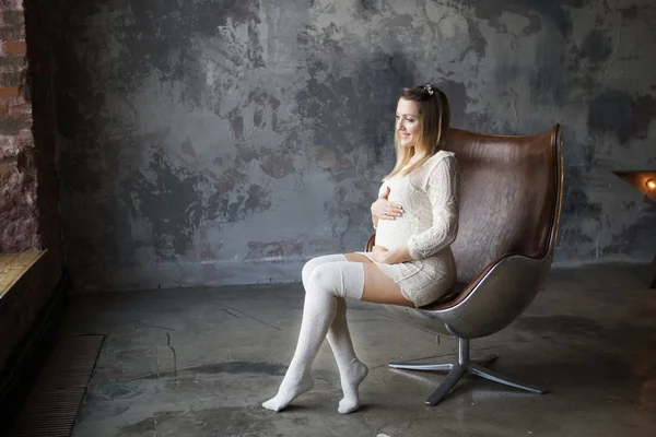 Femme enceinte Blonde avec un beau sourire dans une robe légère et des golfs blancs assis dans une chaise loft marron près d'une grande fenêtre. En arrière-plan un mur gris — Photo