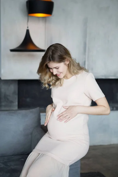 妊娠中のブロンドの女性は、ロフトの奥にベージュのドレスの灰色のソファーに座っています。笑顔と妊娠中の女性が妊娠中の腹で塗抹標本します。 — ストック写真