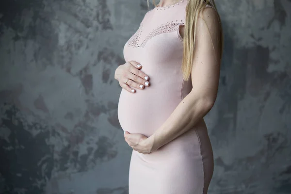 Mulher loira grávida em um vestido leve abraçando sua barriga. No fundo uma parede cinza — Fotografia de Stock