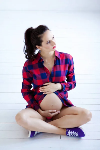 Een zwangere vrouw zit op een witte houten vloer. Paarse sneakers en een rood shirt in een kooi — Stockfoto