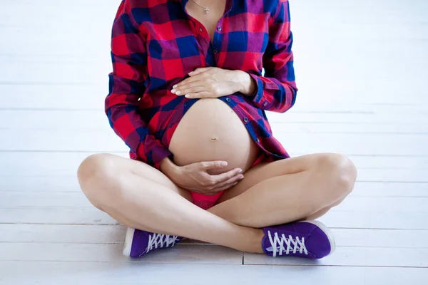 Een zwangere vrouw zit op een witte houten vloer. Paarse sneakers en een rood shirt in een kooi — Stockfoto