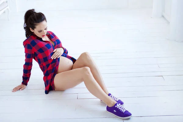 Een zwangere vrouw zit op een witte houten vloer. Paarse sneakers en een rood shirt in een kooi — Stockfoto