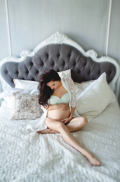 Terhes nő egy szép fehér peignoir és fehérnemű. Terhes nő könnyű nagy ágyon, forgatás felülről — Stock Fotó