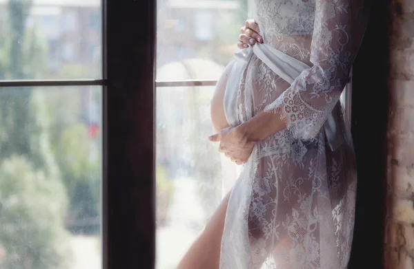 Mujer embarazada en un hermoso peignoir blanco y lencería. Una mujer embarazada está parada junto a la ventana —  Fotos de Stock
