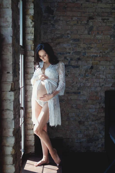 Mulher grávida em um belo peignoir branco e lingerie. Uma mulher grávida está junto à janela. — Fotografia de Stock