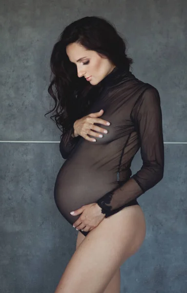 W ciąży brunetka w przejrzyste ciało czarne objął jej ciąży brzuch i spojrzał na niego. Szare tło — Zdjęcie stockowe