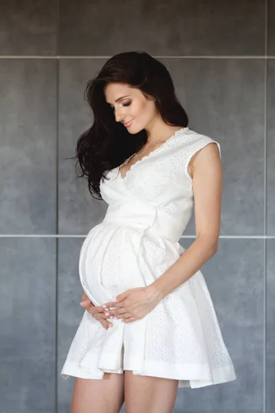 W ciąży brunetka w białej sukni objął jej ciąży brzuch i spojrzał na niego. Szare tło — Zdjęcie stockowe