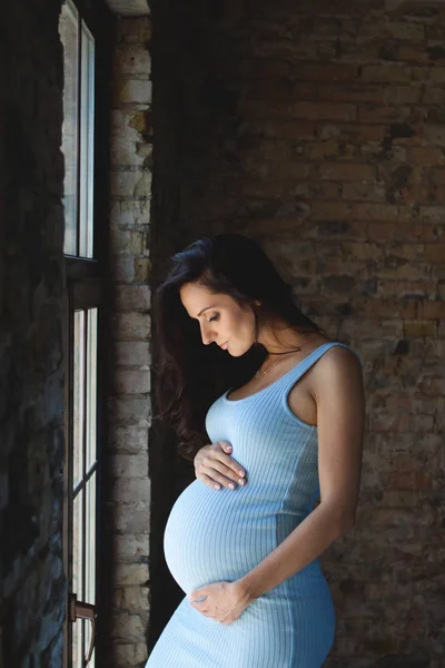 Kobiety w ciąży brunetka w niebieskiej sukience przytulił jej brzuch w ciąży. W ciąży kobieta stoi przy oknie — Zdjęcie stockowe