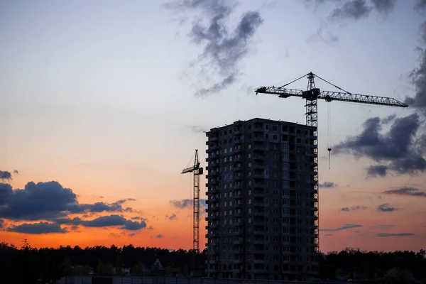 Guindaste e canteiro de obras contra o pôr do sol. Construção de edifício inacabado e guindastes de construção contra o fundo do céu azul. Conceito de desenvolvimento urbano Imagens Royalty-Free