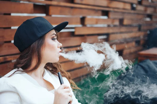 Young woman in a black cap smoking a hookah outdoors. The pleasure of smoking. Background a wooden wall — Stock Photo, Image