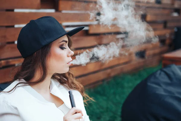 Young woman in a black cap smoking a hookah outdoors. The pleasure of smoking. Background a wooden wall — Stock Photo, Image