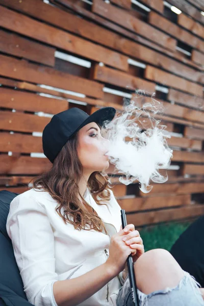 Young woman in a black cap smoking a hookah outdoors. The pleasure of smoking. Background a wooden wall — Stock Photo, Image