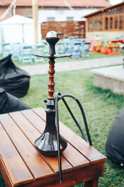 A black hookah is on a wooden table outside. Background lifestyle concept. Background black pouffes.  Hookah in the open air in the summer. — Stock Photo, Image