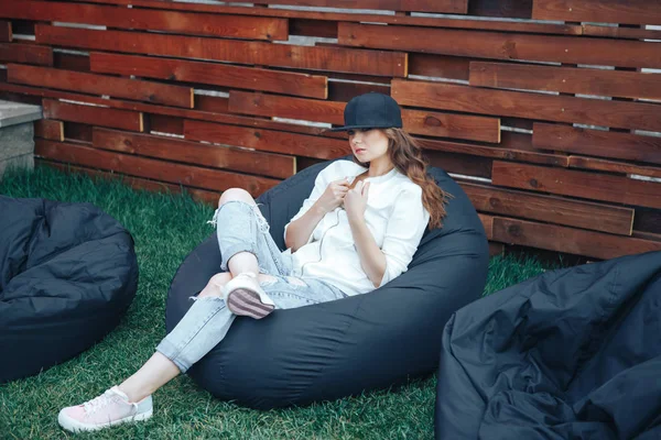 Mujer joven con una gorra negra se sienta en una bocanada negra al aire libre. Bolsa de frijol negro silla al aire libre Fotos De Stock Sin Royalties Gratis