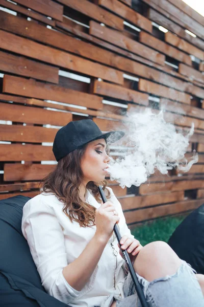 Young woman in a black cap smoking a hookah outdoors. The pleasure of smoking. Background a wooden wall — Stock Photo, Image