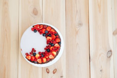 Beyaz peynirli kremalı pasta ganache ve kırmızı meyveler beyaz bir stand ile dekore edilmiştir. Pastayı ahşap bir masa üzerinde duruyor. Üstten görünüm