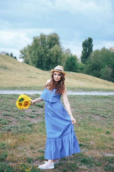 Jovem mulher em um chapéu de palha em um parque com girassóis — Fotografia de Stock