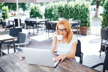 Kızıl saçlı kadın kahve içme ve yaz kafede dizüstü bakarak. Genç bir kadın oturuyor bir kafe ve işleri