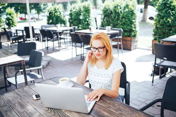 Joven mujer pelirroja mirando a la computadora portátil en el café de verano. Una mujer joven se sienta en un café y trabaja detrás de una computadora portátil — Foto de Stock