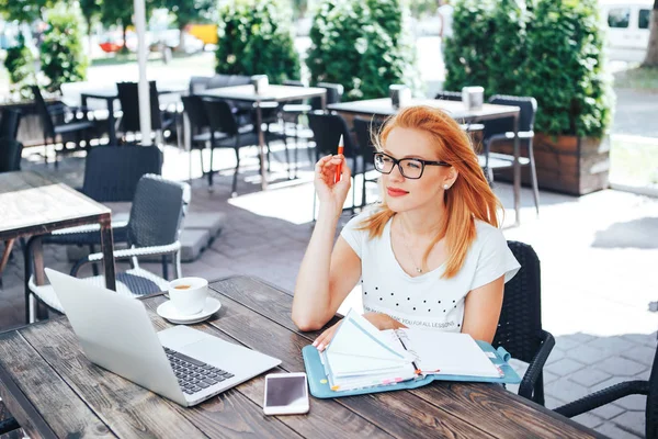 Joven pelirroja mujer piensa qué escribir en un cuaderno en un café de verano. Una joven se sienta en un café y trabaja — Foto de Stock