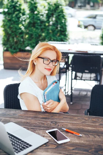 Una joven pelirroja sosteniendo un cuaderno azul en un café de verano. Una joven se sienta en un café y trabaja — Foto de Stock