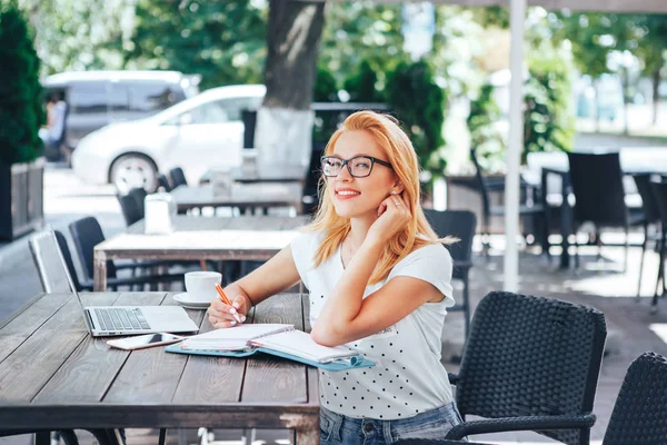 Joven mujer pelirroja sonriendo sentada en el café de verano. Una mujer joven se sienta en un café y trabaja detrás de una computadora portátil — Foto de Stock