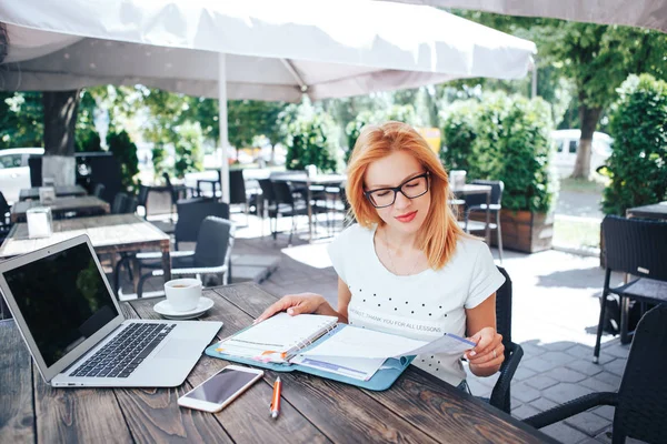 Una joven pelirroja mirando en un cuaderno en un café de verano. Una joven se sienta en un café y trabaja — Foto de Stock