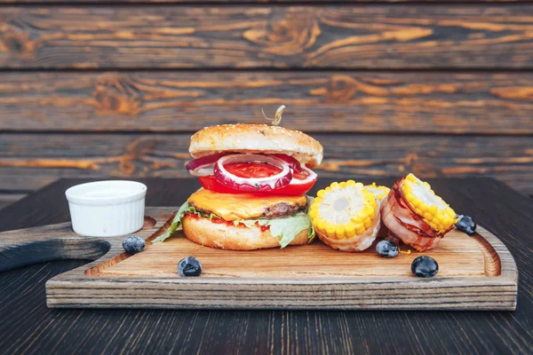 Deliciosa hamburguesa casera a la parrilla con carne de res, cebolla, tomate, queso, ensalada y maíz. Hamburguesas BBQ sobre tabla de cortar sobre fondo de madera — Foto de Stock