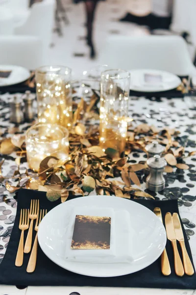 La mesa de la cena está decorada en negro y oro. Guirnalda en un frasco de vidrio. Tenedor y cuchillo dorado Fotos De Stock Sin Royalties Gratis