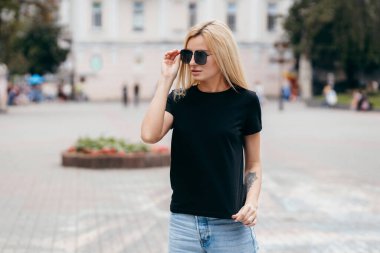 Siyah tişörtlü, gözlüklü şık sarışın kız sokak ortasında poz veriyor. Sokak fotoğrafı.