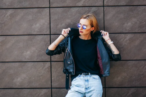 Blonde girl wearing black t-shirt, glasses and leather jacket posing against street , urban clothing style. Street photography