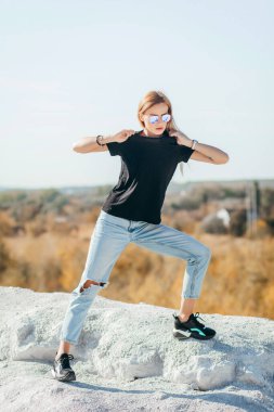 Siyah tişörtlü ve gözlüklü tipik sarışın kız sokak ortasında poz veriyor, kentsel giyim tarzı. Sokak fotoğrafı.