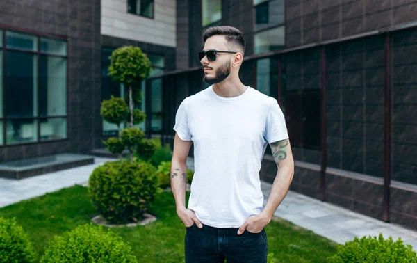 A young stylish man with a beard in a white T-shirt and glasses. Street photo