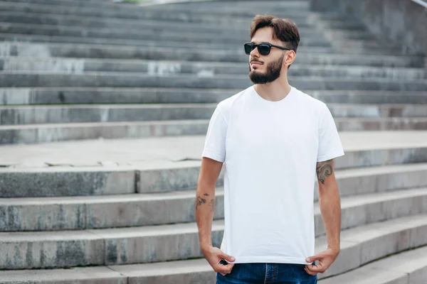 A young stylish man with a beard in a white T-shirt and glasses. Street photo