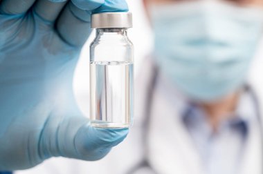 Doktorun eli beyaz arka planda mavi eldivenler giyiyor ve elinde Covid-19 aşısı var..