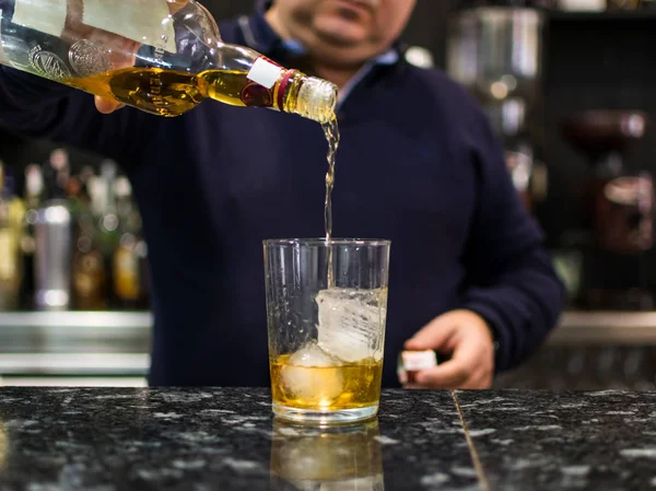 Barman pouring whiskey in front of whiskey glass and bottles on black table with reflection