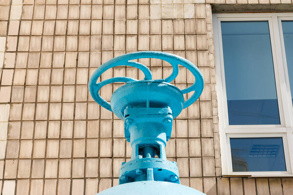 big blue valve on the gray tile wall with window background. A valve for controlling the output of liquids, steam or gas in certain mechanisms