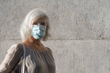 Gri saçlı, orta yaşlı bir kadına yakın çekim. Sokakta dikilen, kovid19, virüs ve hastalıktan korunmak için yüz maskesi takan bir kadın.