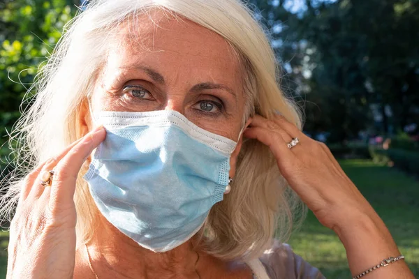 beautiful gray-haired middle aged woman straightens her protective mask outdoors. woman wearing face mask outside in the Corona virus COVID-19 pandemic