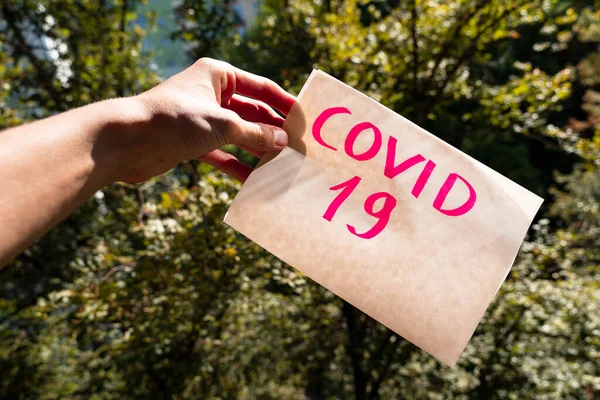 covid-19 lettering on a piece of paper that hand is holding, blurred green trees in a background, outdoors. second wave of coronavirus infection concept