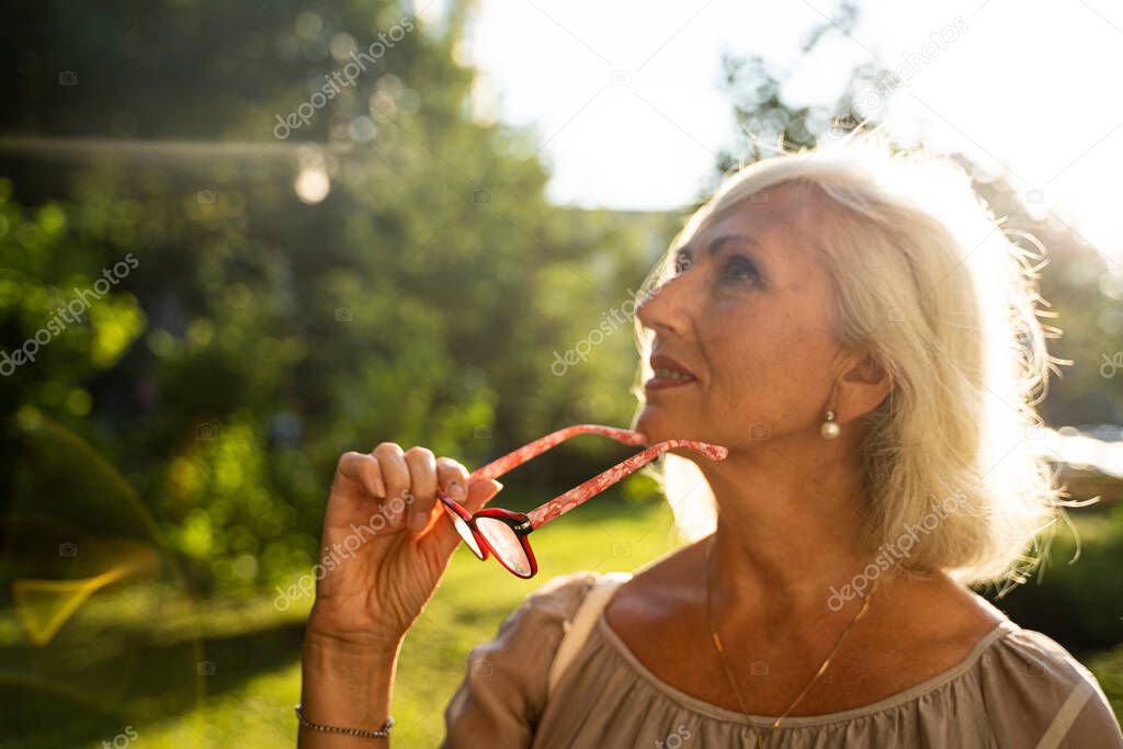 blurred portrait of pensive gray-haired woman takes off her glasses and looks up while on the street in summer