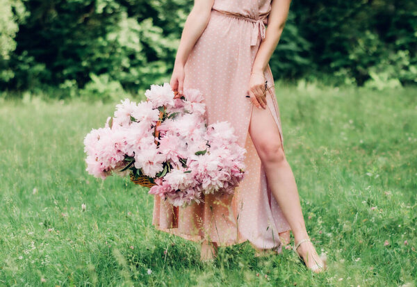 A girl in a pink dress in peas on a green background holds in one hand a basket with pink peonies and in the other glasses with a gold rim
