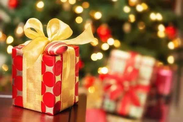 红色和金色的圣诞礼物 桌子上有一个弓和一个装饰品 节日期间背景与圣诞树灯和礼物在树之下 — 图库照片