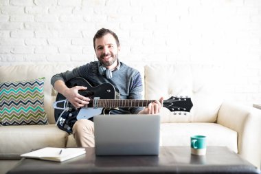 Kanepe ile dizüstü bilgisayar önünde oturup gitar ile mutlu genç adam portresi